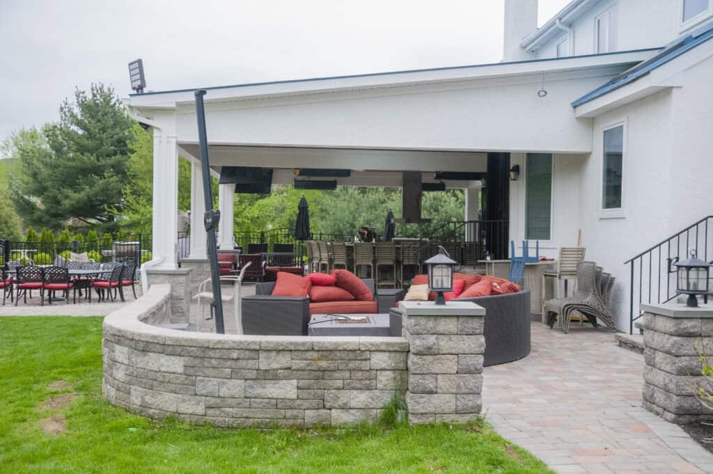 A spacious backyard patio with outdoor seating, red cushions, a dining table, and a covered area, surrounded by stone walls and greenery.
