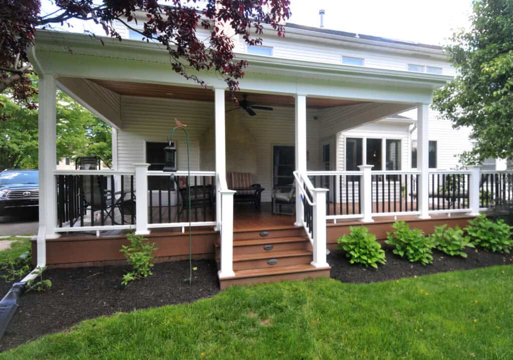 Covered porch attached to a white house, with wooden steps, white railings, ceiling fan, seating area, and green shrubs planted along the edge, overlooking a grassy yard.