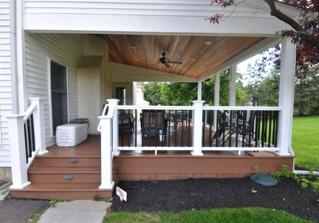A covered backyard deck with brown steps and flooring, white railing with black balusters, outdoor seating, a ceiling fan, and a grassy yard in the background. A white cooler sits near the door.