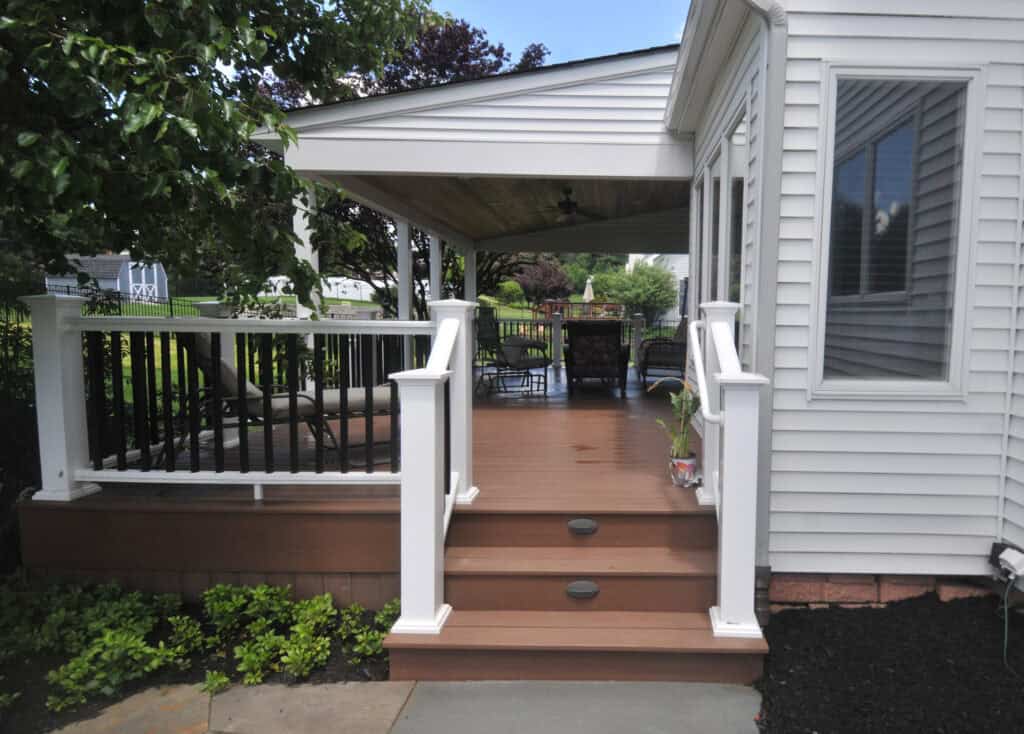 A wooden deck with white railings and steps leads to a covered porch attached to a white house. Patio furniture is visible under the roof, surrounded by greenery and a tree to the left.