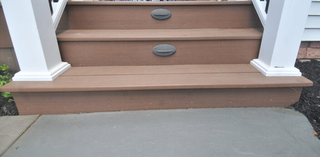 Wooden porch steps with brown treads and white rail posts, viewed from the bottom. There are two oval-shaped step lights mounted on the risers. The steps lead from a concrete surface to a higher porch.