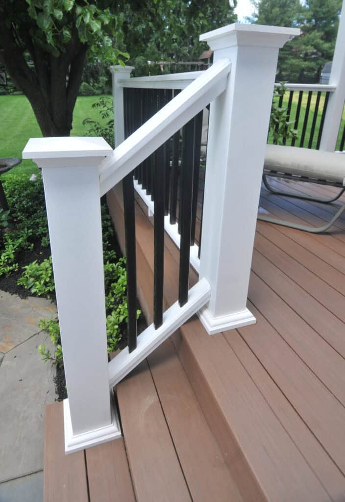 White and black deck railing on brown composite steps, with greenery and a tree in the background. The railing posts are white with black vertical balusters and a landscaped yard nearby.