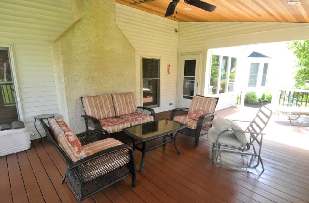A covered patio with wicker furniture, including a glass-top coffee table, cushioned chairs, a love seat, a rocking chair, and a chimney; the floor is wood, and large windows overlook a green yard.