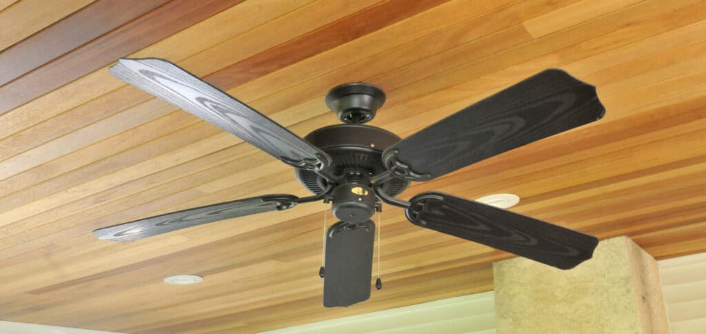 A black ceiling fan with five blades is mounted on a wooden paneled ceiling, with part of a beige wall visible in the background.
