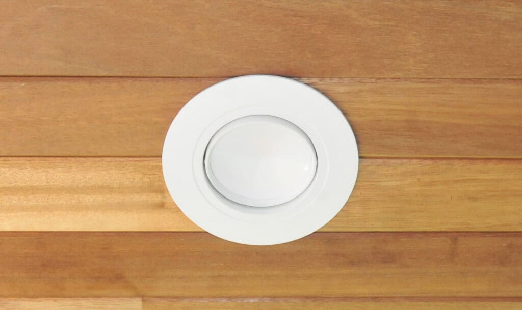 A round, white recessed ceiling light fixture is set into a wooden ceiling with horizontal planks. The light is off, and the wood has a warm, natural finish.
