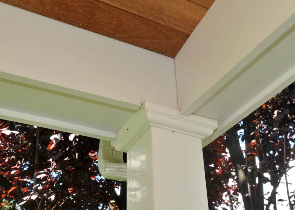 Close-up view of a porch corner showing neatly joined white wooden trim, columns, and molding beneath a wood-paneled ceiling, with leafy trees visible in the background.