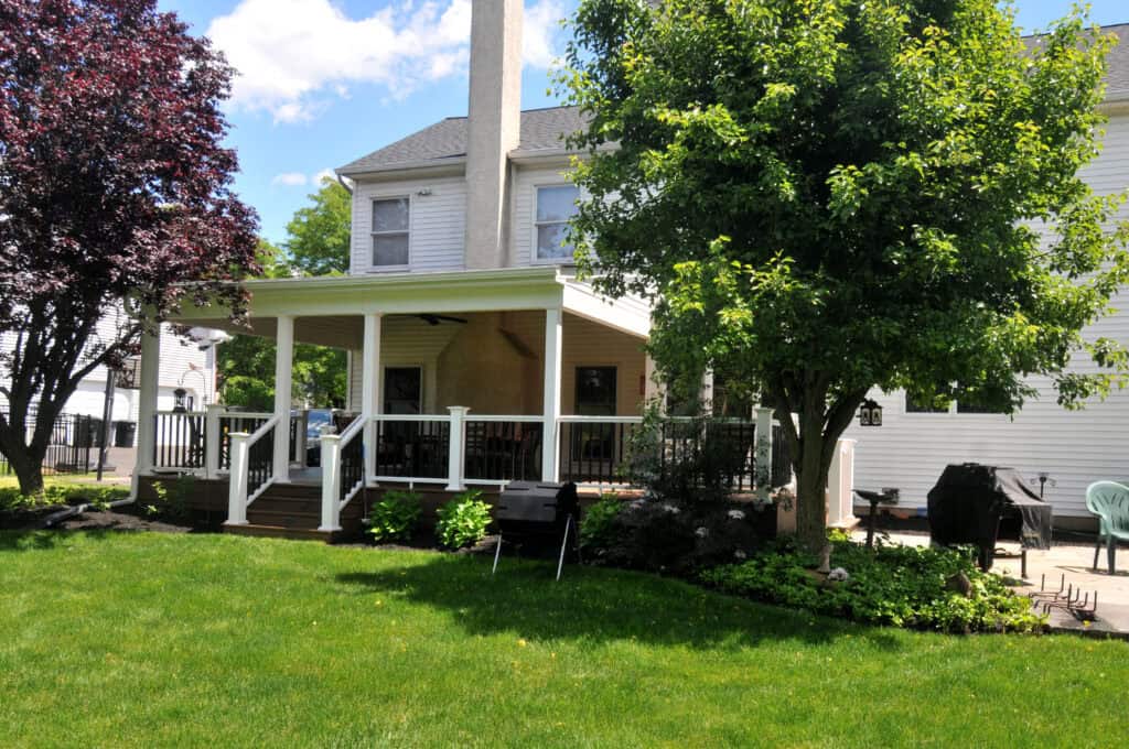A backyard view of a house with a covered wooden deck, outdoor furniture, a grill, lush green lawn, and mature trees providing shade on a sunny day.