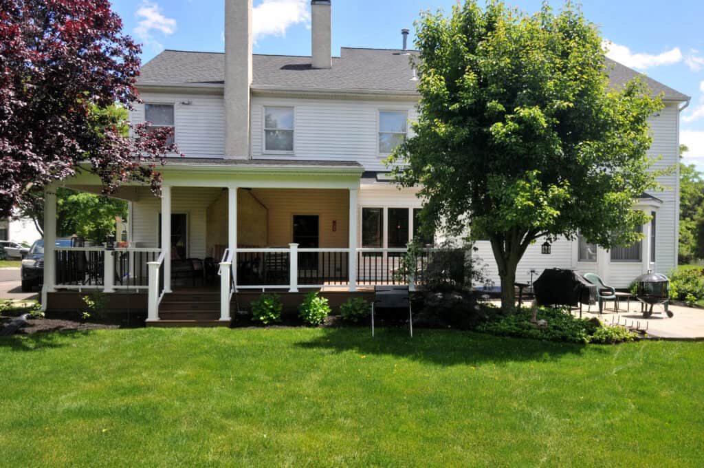 A two-story white house with a covered back porch, outdoor furniture, a grill, and a green lawn with trees and shrubs on a sunny day.