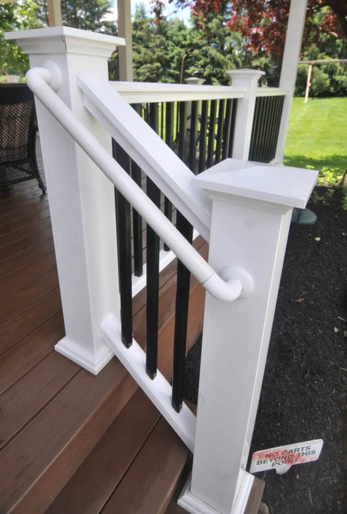 White wooden porch railing with black vertical balusters and a matching handrail on brown wooden steps. In the background, there is a grassy yard and trees. A small sign on the ground says NO CARTS BEYOND THIS POINT.
