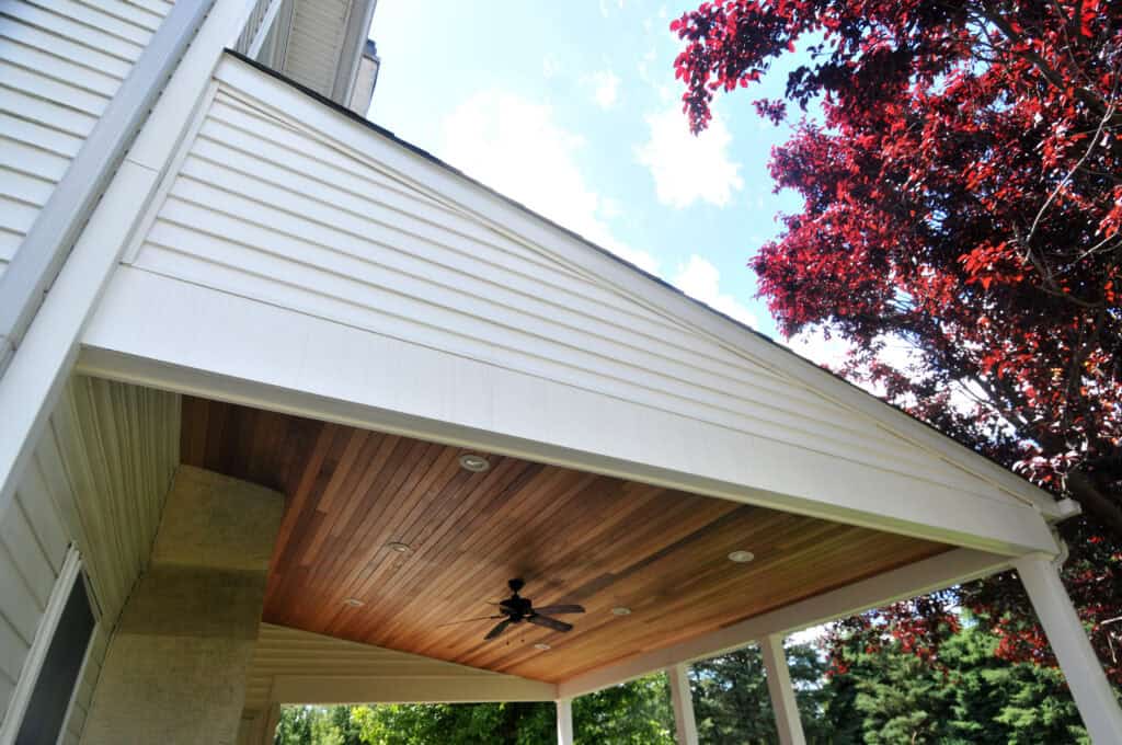 View from below a covered porch attached to a house, featuring a wood-paneled ceiling with recessed lights and a ceiling fan. A red-leaved tree is visible on the right, and the sky is partly cloudy.