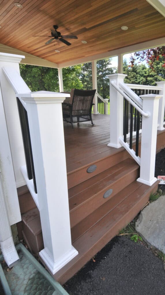 A covered wooden porch with brown steps, white railings with black balusters, and a ceiling fan; a black chair and table are visible on the porch, surrounded by green trees and grass.