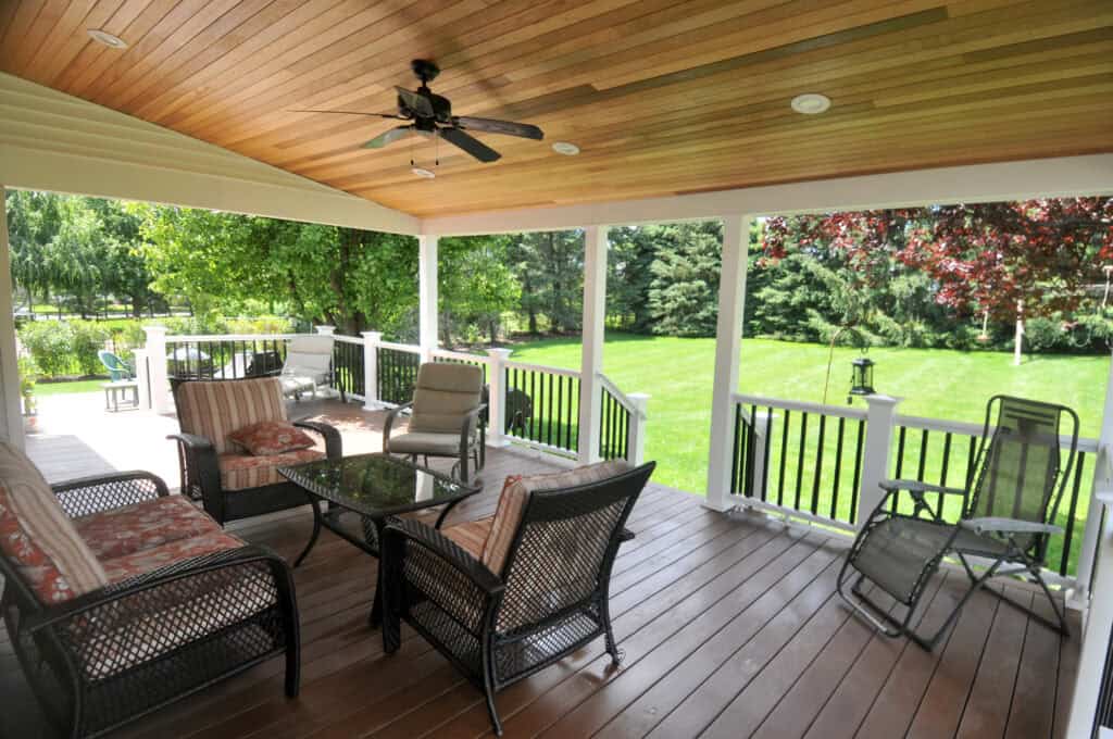 A covered porch with wicker furniture, cushioned chairs, a glass coffee table, and a ceiling fan overlooks a green backyard with trees and a neatly mowed lawn.