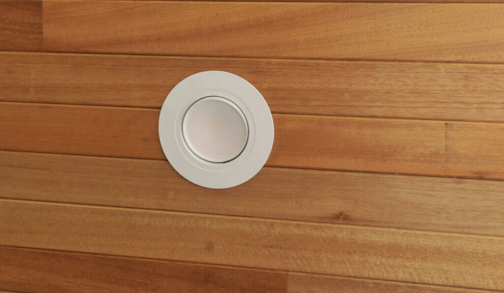 A round, white recessed ceiling light is installed in a wooden plank ceiling. The wood panels are horizontal and light brown in color.