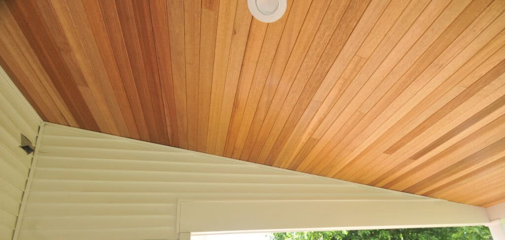 Angled view of a ceiling with horizontal wooden planks, a recessed circular light fixture, and an adjacent sloped cream-colored wall paneling. Some greenery is visible outside near the bottom edge.