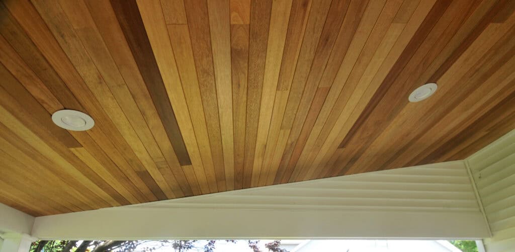 Wood-paneled ceiling with two recessed round lights, featuring slanted beige trim below. Natural light highlights the wood grain, and part of a tree and house roof are visible outside through an opening.