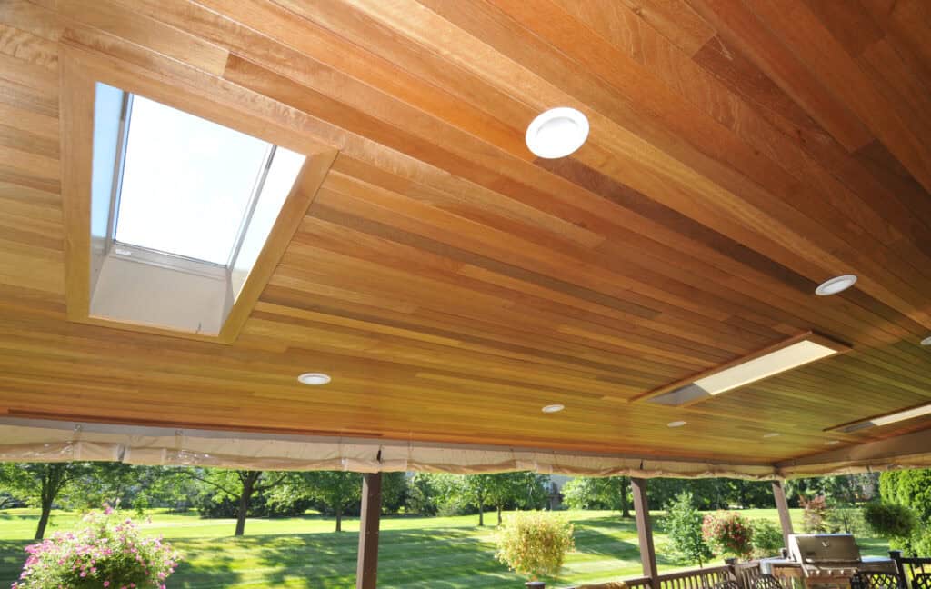 Wood-paneled ceiling with skylights and recessed lights on a covered patio, overlooking a green lawn and trees in the background.