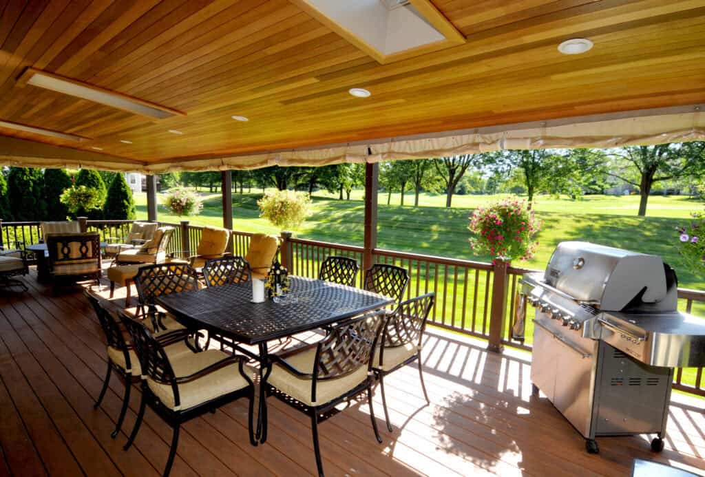 A covered wooden deck with a metal dining table and chairs, cushioned seating, a stainless steel grill, potted flowers, and a view of a grassy yard with trees in the background.