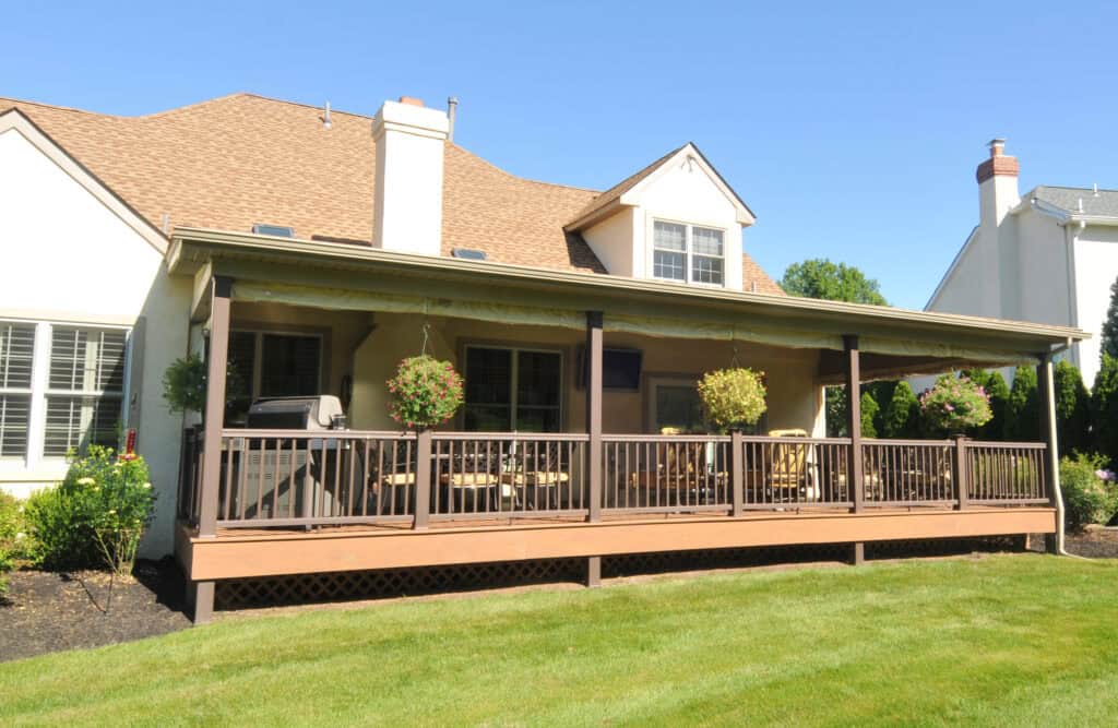 A spacious backyard deck with a roof, railings, patio furniture, hanging flower baskets, and a grill, attached to a light-colored house with a sloped roof on a sunny day.