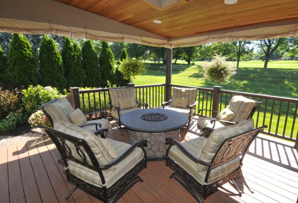 A covered outdoor patio with six cushioned chairs arranged around a circular fire pit table, surrounded by wooden railings and lush green trees and bushes in the background.