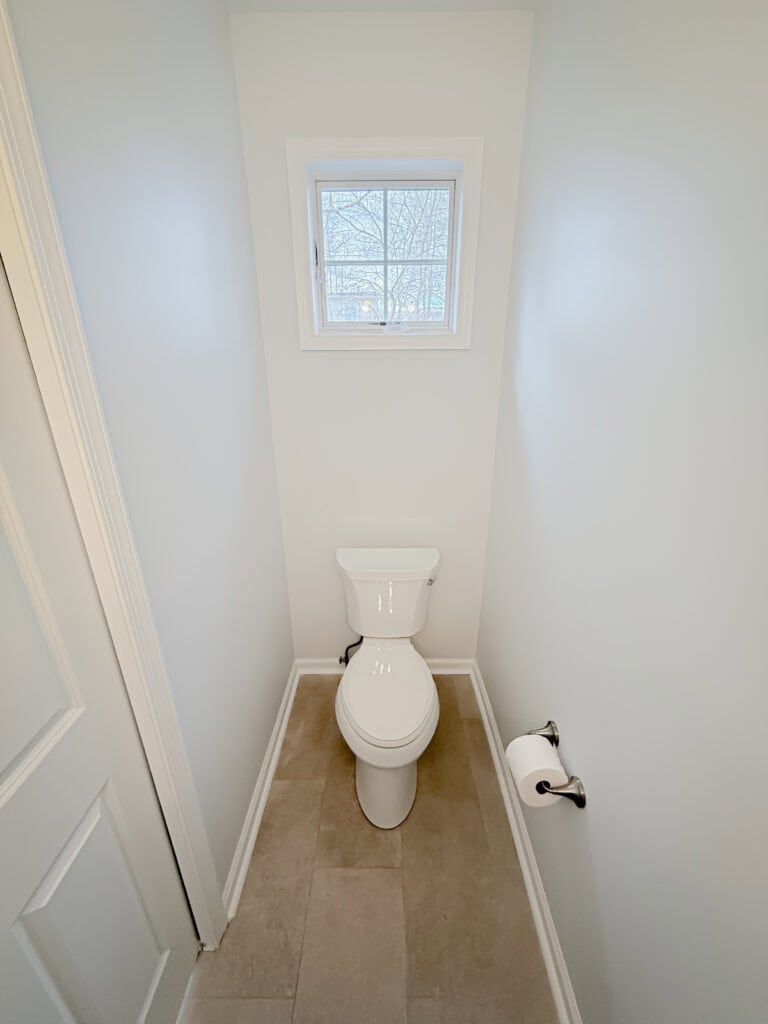 A small, narrow bathroom with a white toilet centered beneath a small window, white walls, beige tiled floor, and a toilet paper holder mounted on the right wall.