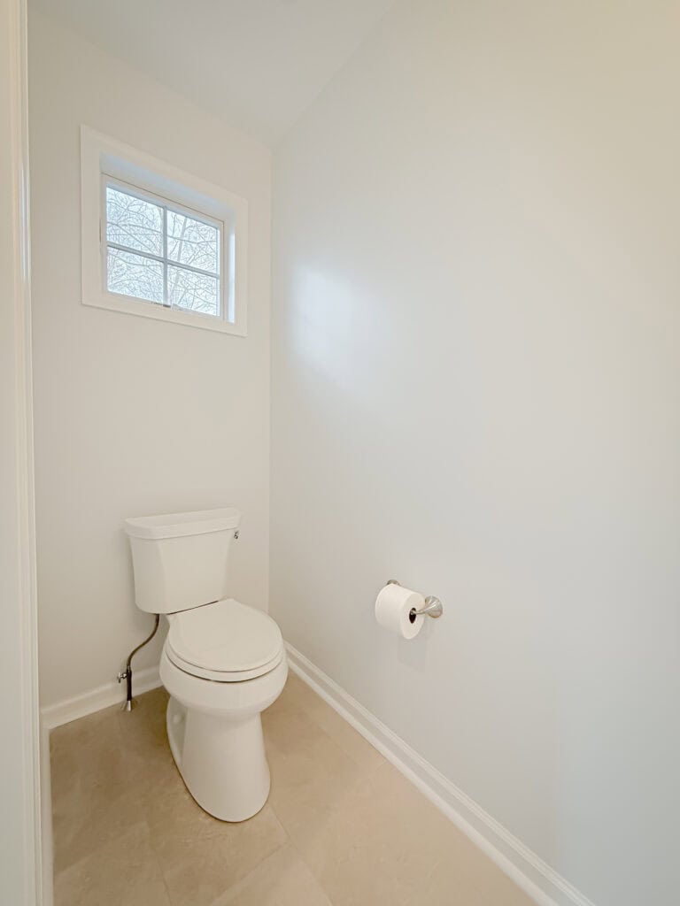 A white toilet with a roll of toilet paper mounted on the wall beside it sits in a small, clean bathroom with cream-colored tile flooring and a window above.