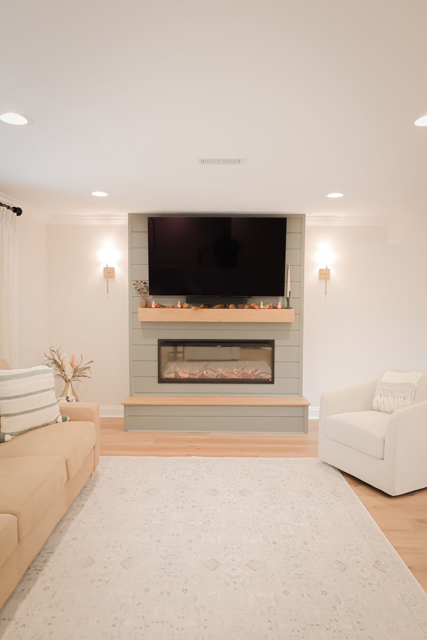 A modern living room with a beige sofa, white armchair, light patterned rug, and a wall-mounted TV above a built-in electric fireplace decorated with small autumn accents. The room has light wood floors and soft lighting.