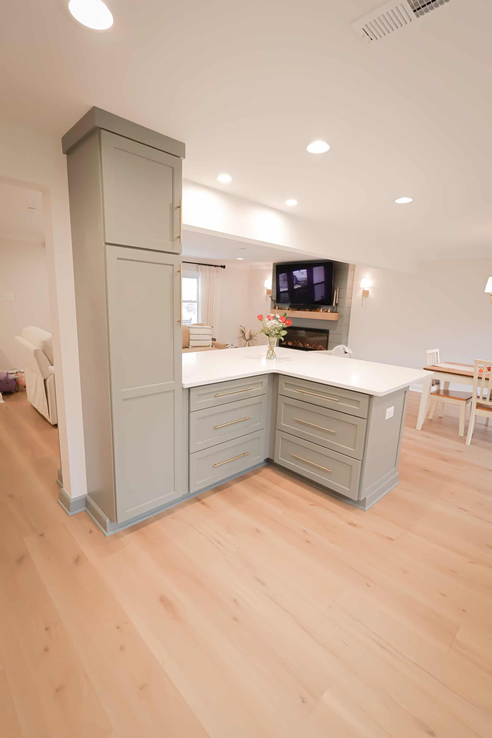 Modern kitchen remodel with light wood flooring, pale green cabinets, and white countertops. A bouquet of flowers sits on the counter. The space opens to a living room with a TV and a dining area with a table and chairs.