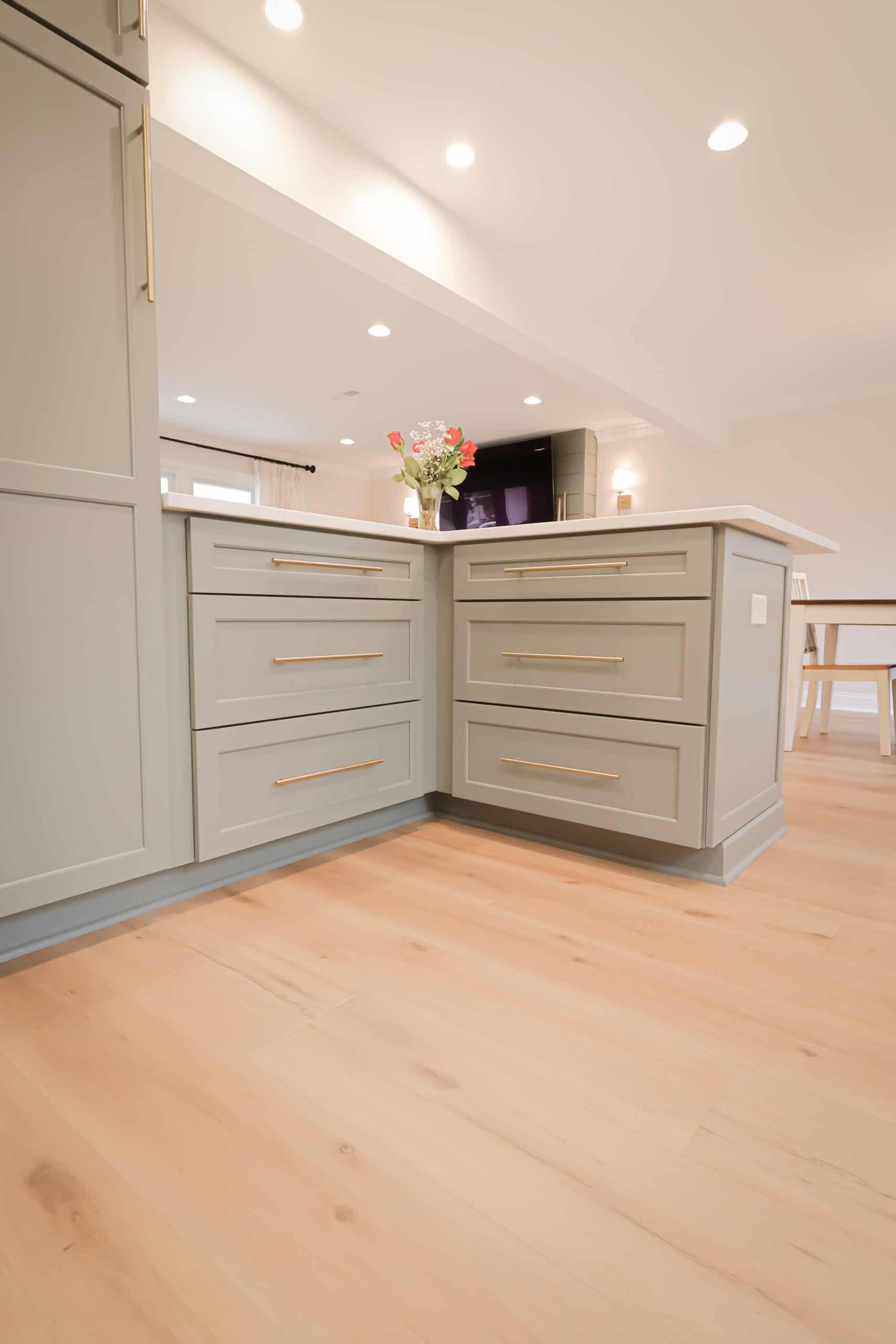 A modern kitchen remodel features light gray cabinets with long gold handles, light wood flooring, and a vase of flowers on the countertop under recessed ceiling lights.