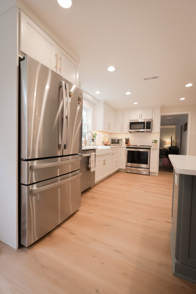 Modern kitchen remodel with light wood floors, white cabinets, stainless steel appliances including a refrigerator, stove, and microwave, and bright overhead lighting. A window above the sink brings in natural light.