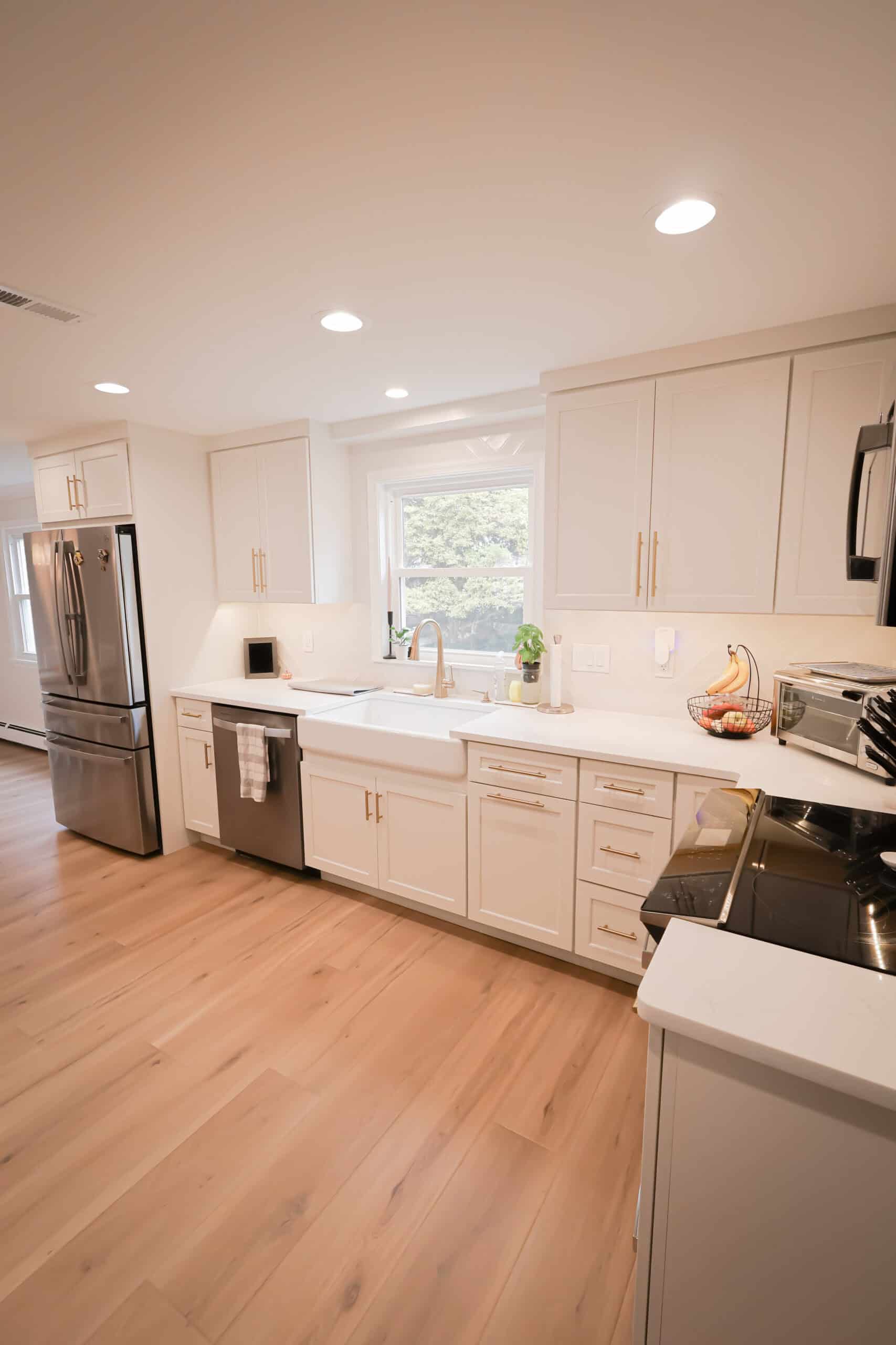 A modern kitchen remodel with white cabinets, stainless steel appliances, a farmhouse sink under a window, and light wood flooring. A fruit basket and small plant sit on the white countertop, completing this inviting space.
