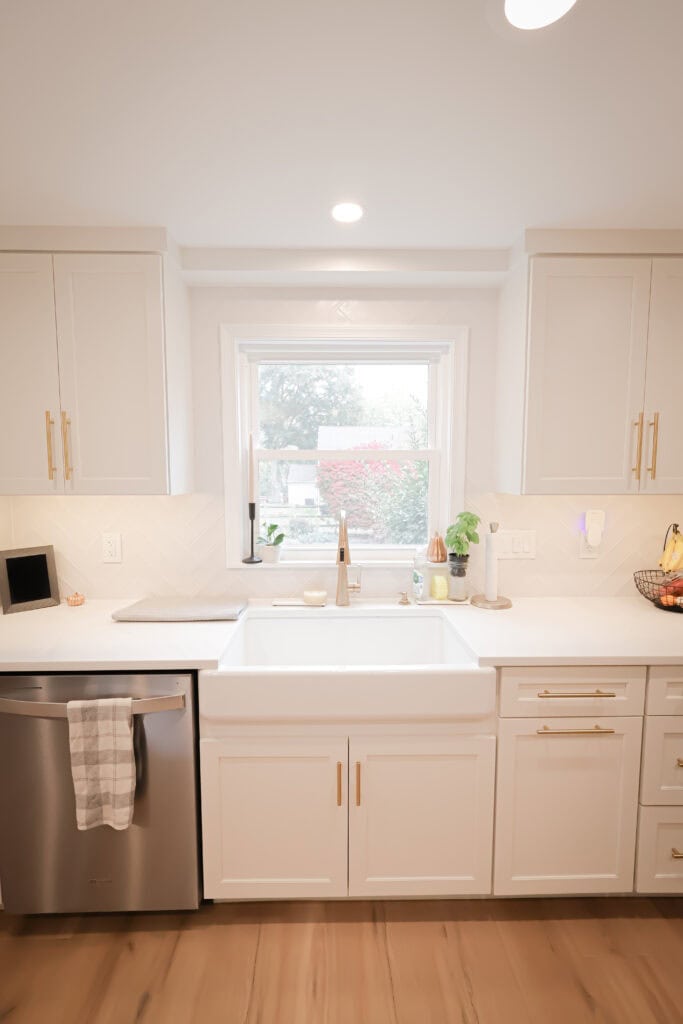Bright, modern kitchen remodel with white cabinets, gold handles, a farmhouse sink, and stainless steel dishwasher. A window above the sink lets in natural light. Countertops hold a plant, soap dispenser, fruit basket, and decor.