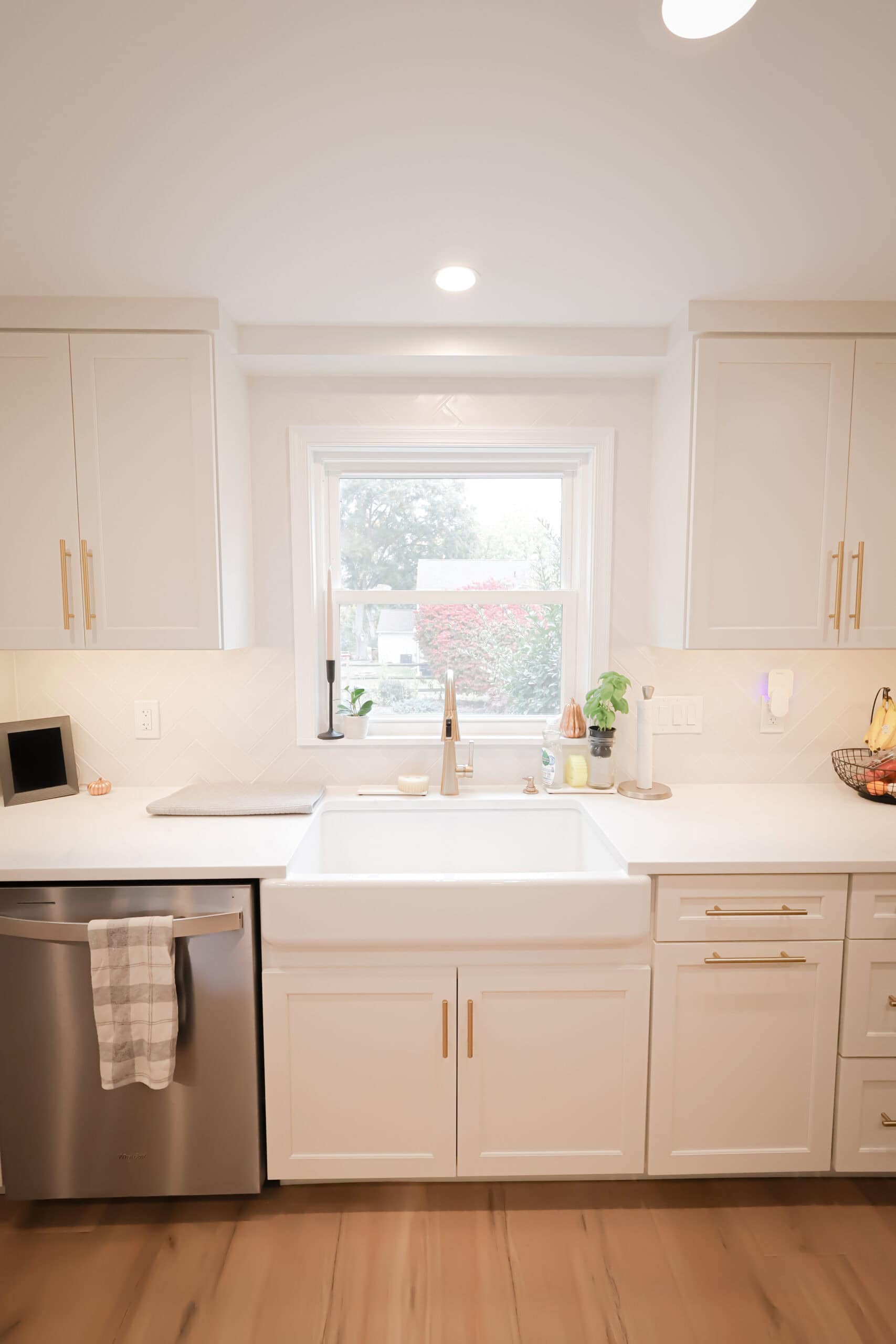 Bright, modern kitchen remodel with white cabinets, gold handles, a farmhouse sink, and stainless steel dishwasher. A window above the sink lets in natural light. Countertops hold a plant, soap dispenser, fruit basket, and decor.