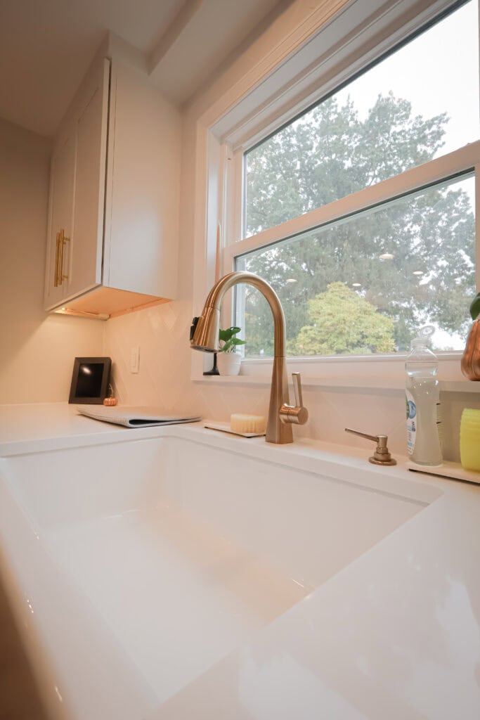 Modern kitchen sink with a gold faucet, white countertop, dish soap, and a yellow sponge—perfect inspiration for kitchen remodels. A large window frames green trees outside, while cabinets with gold handles sit on the left.