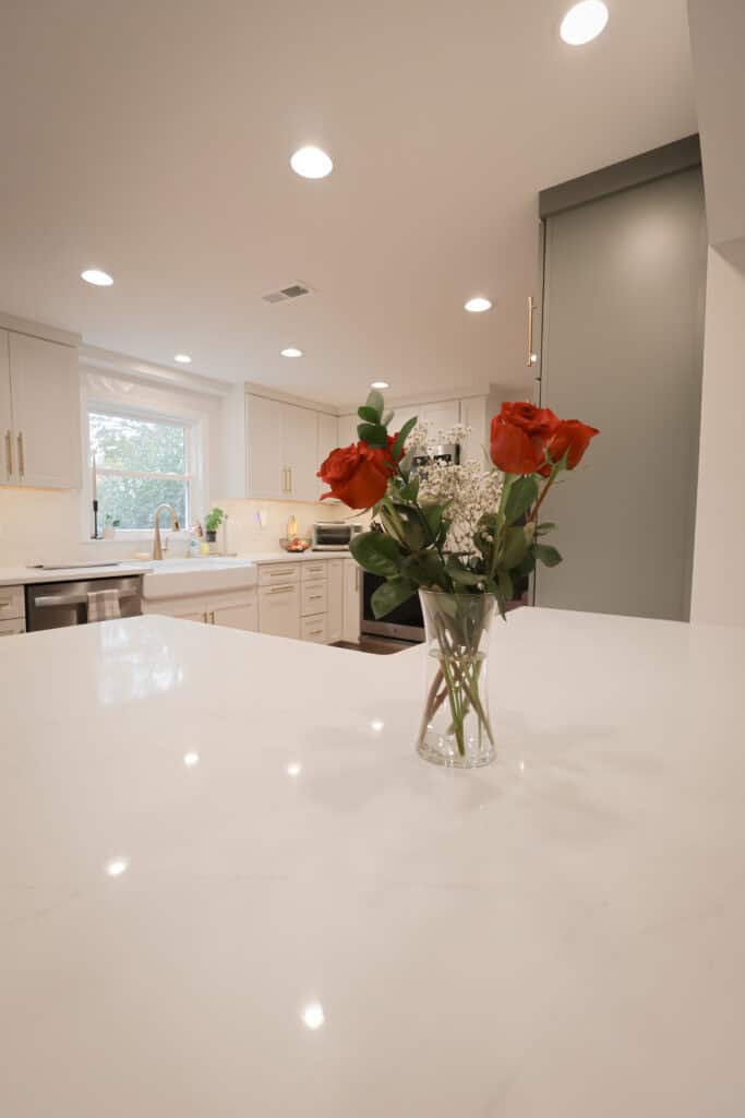 A vase with red roses and white baby&#039;s breath sits on a white marble kitchen island, highlighting the elegance of recent kitchen remodels featuring white cabinets, a farmhouse sink, stainless steel appliances, and recessed lighting.