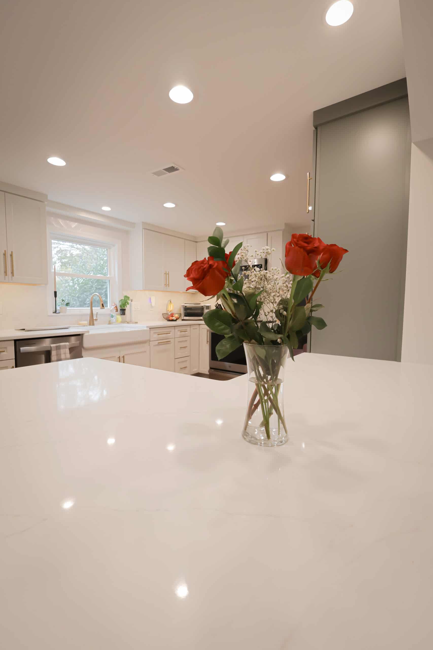 A vase with red roses and white baby's breath sits on a white marble kitchen island, highlighting the elegance of recent kitchen remodels featuring white cabinets, a farmhouse sink, stainless steel appliances, and recessed lighting.