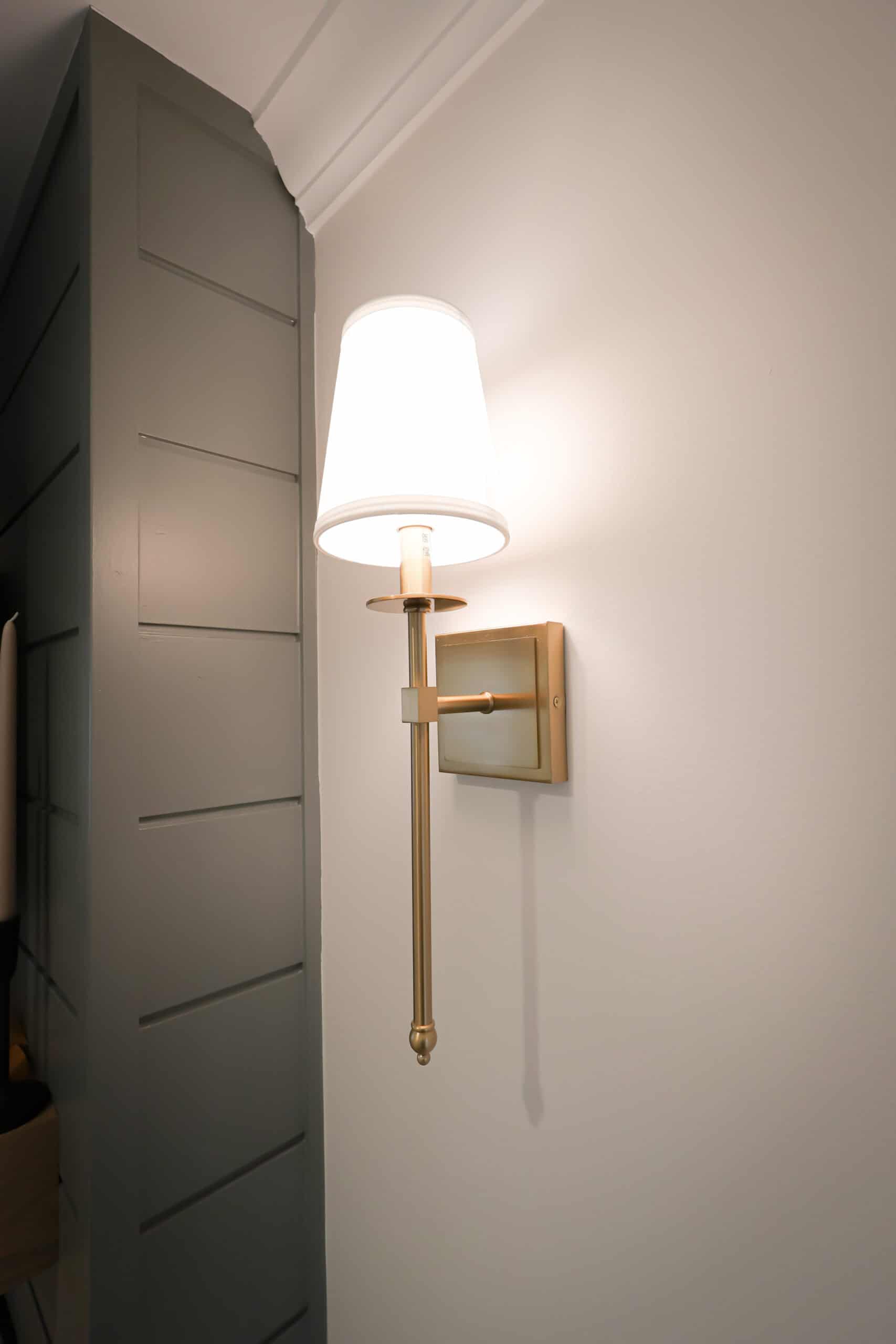 A modern wall-mounted brass sconce with a white lampshade glows against a white wall, next to a gray shiplap accent—perfect for adding sophisticated lighting to kitchen remodels.