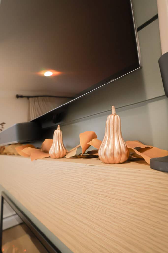 Two decorative metallic pumpkins and dried leaves sit on a light wooden shelf beneath a mounted flat-screen TV, adding an autumn touch—perfect inspiration for cozy kitchen remodels this season.