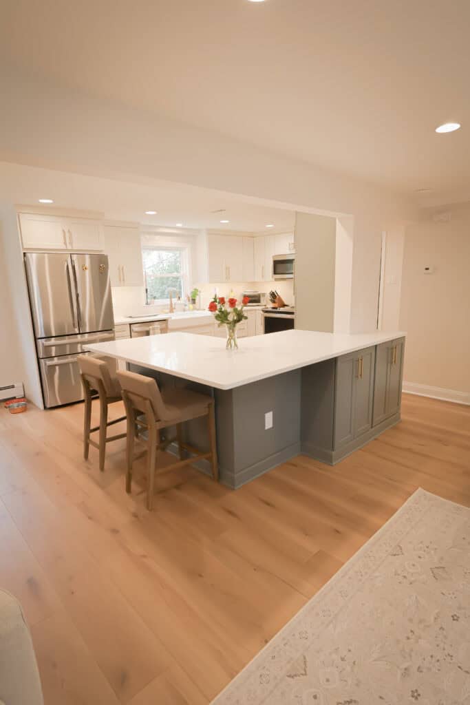 Modern kitchen remodel with light wood floors, white cabinets, stainless steel appliances, and a large island featuring a white countertop, gray base, and two beige barstools. A vase with flowers sits on the island.