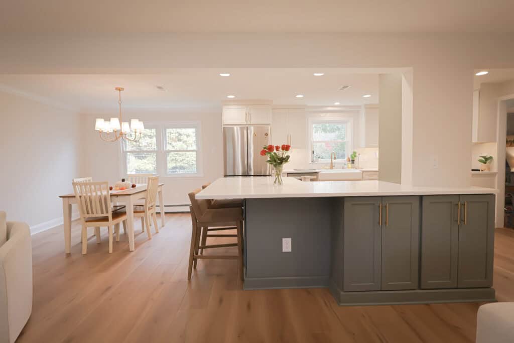 A modern kitchen remodel features a large island, gray cabinets, and bar stools, open to a dining area with a wooden table and white chairs. Natural light streams in from multiple windows, highlighting the light wood floors.