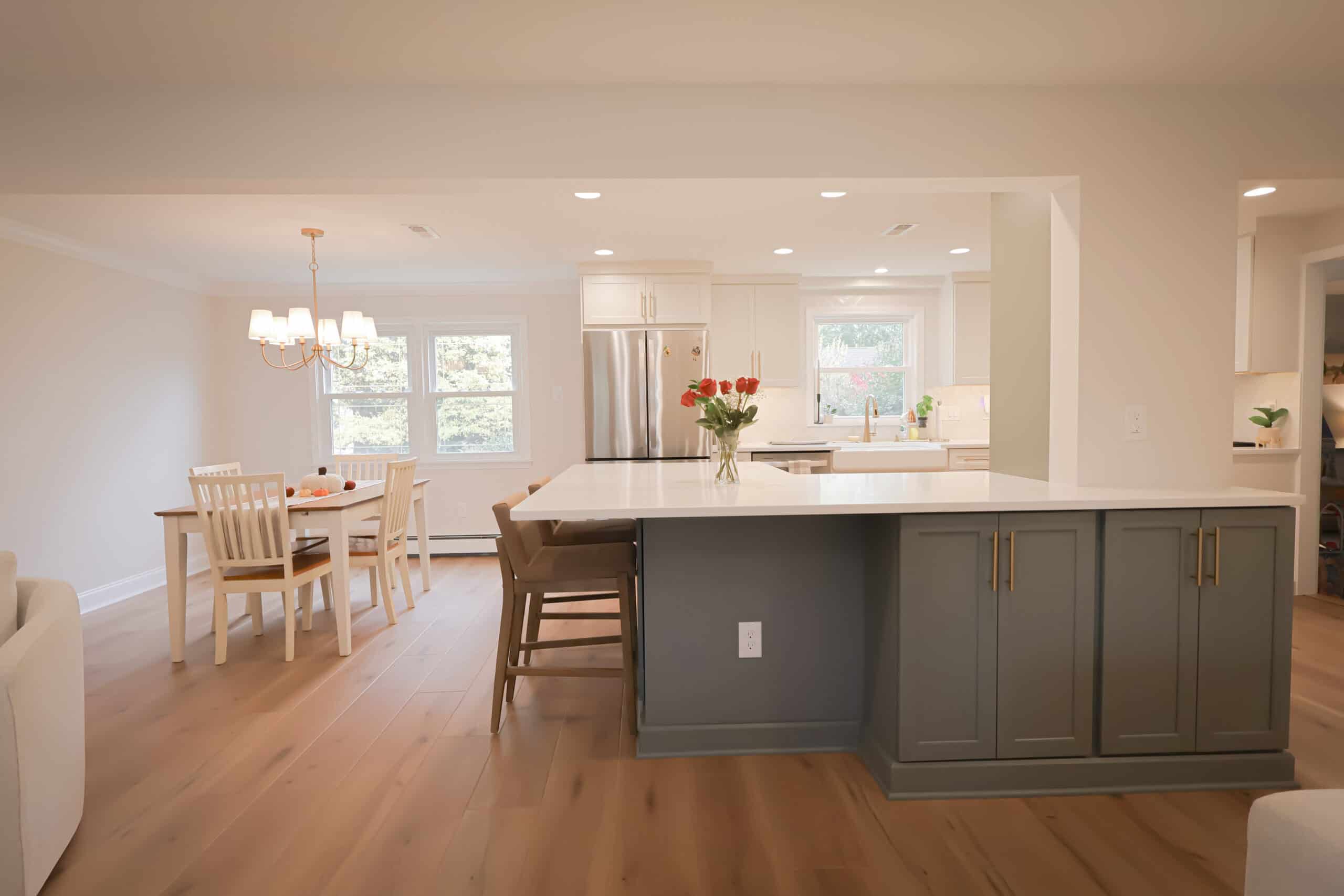 A modern kitchen remodel features a large island, gray cabinets, and bar stools, open to a dining area with a wooden table and white chairs. Natural light streams in from multiple windows, highlighting the light wood floors.