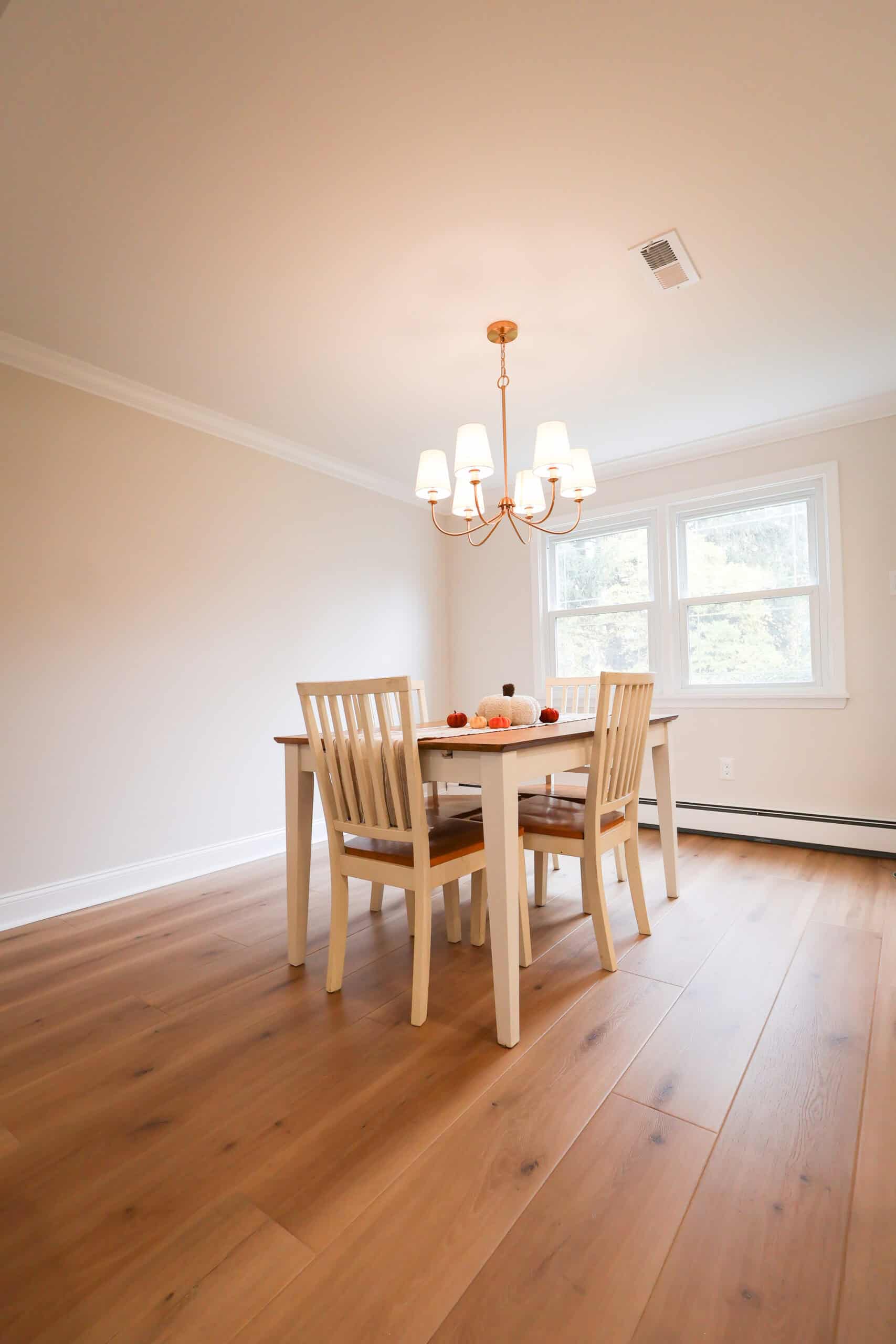 A bright dining room with light wooden floors, a white table, and four matching chairs showcases kitchen remodels at their finest. A chandelier hangs above the table adorned with small pumpkins as sunlight streams in through two windows.