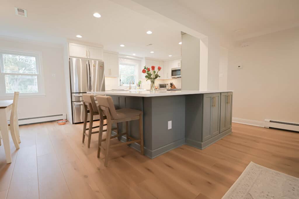 Modern kitchen remodel with gray island, white countertops, three beige bar stools, stainless steel appliances, light wood flooring, and a vase of red roses on the island. Natural light streams in from large windows.