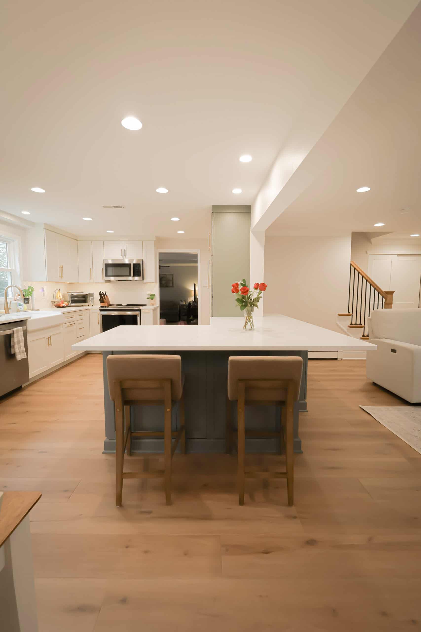 Modern kitchen remodel with light wood floors, white cabinets, a spacious island with two barstools, stainless steel appliances, fresh flowers in a vase, and an open layout that flows to the living area and staircase.