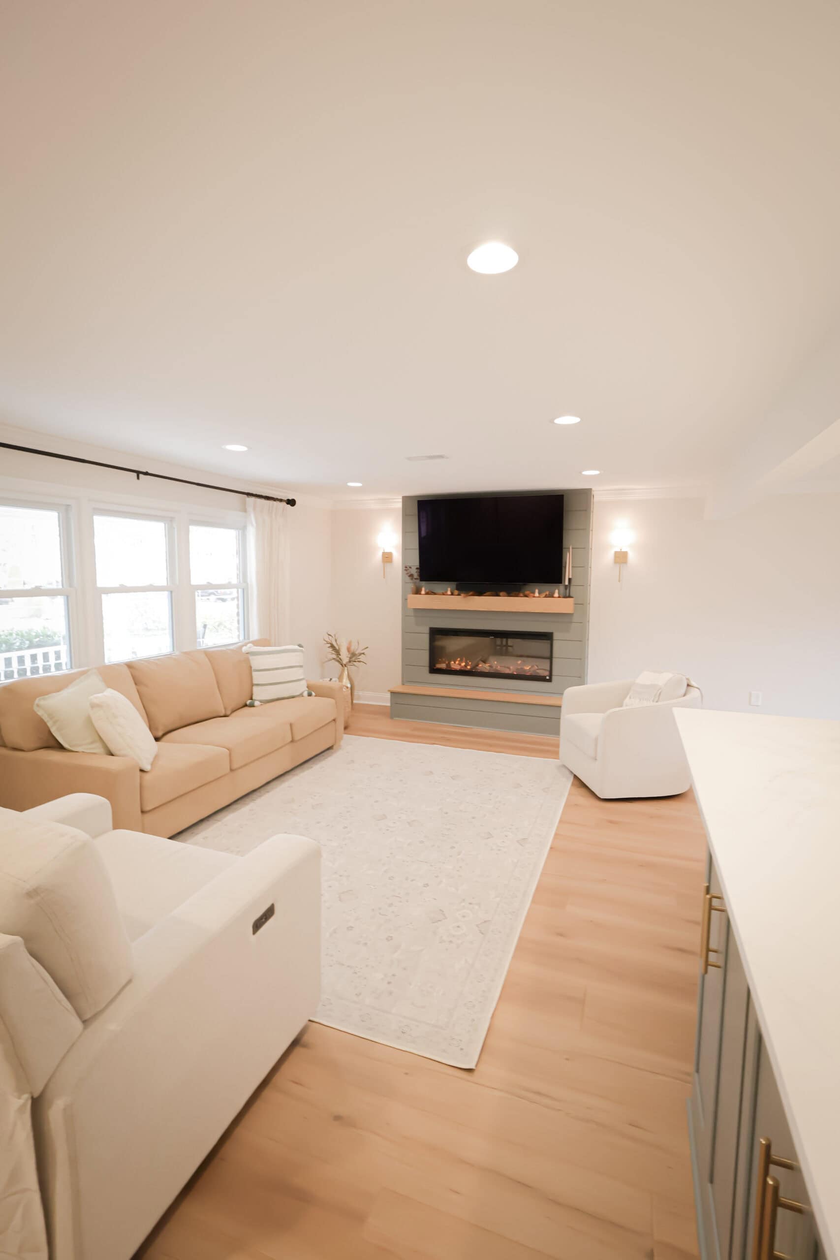 A bright, modern living room with light wood floors, a beige sofa, a white armchair, a large area rug, a wall-mounted TV above a sleek fireplace, and large windows letting in natural light.
