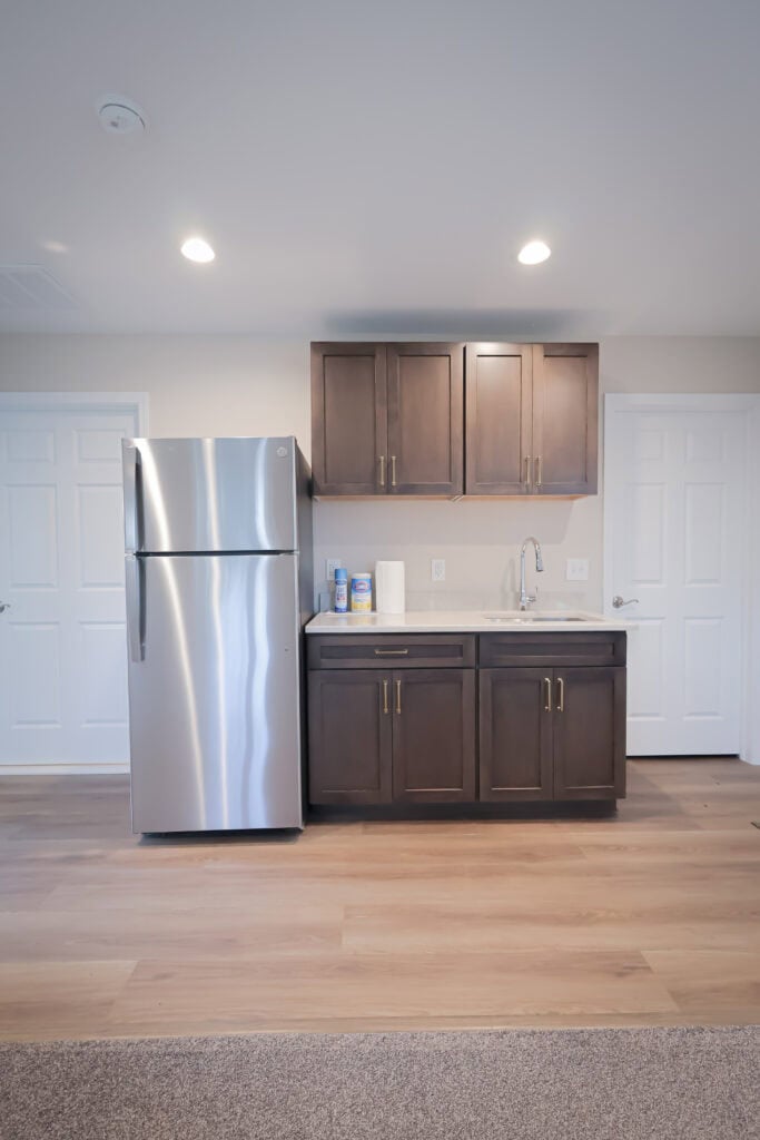 A small kitchen area with a stainless steel refrigerator, dark wood cabinets, a sink, paper towels, and cleaning supplies on the counter. The space has light-colored walls and wood flooring.