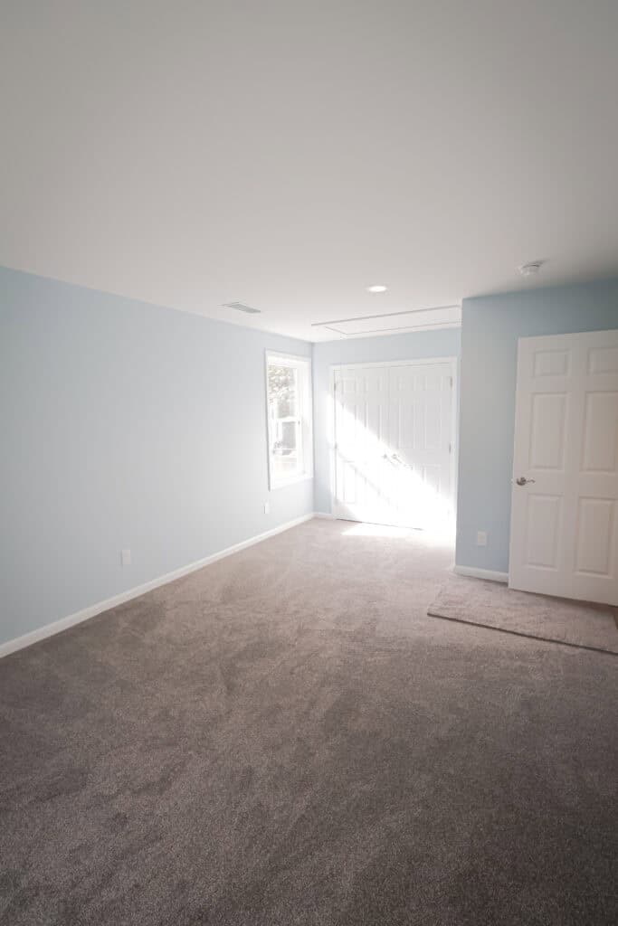 A bright, empty room with light blue walls, gray carpet, a window, and two white doors, one letting in natural sunlight. The space is clean and uncluttered.