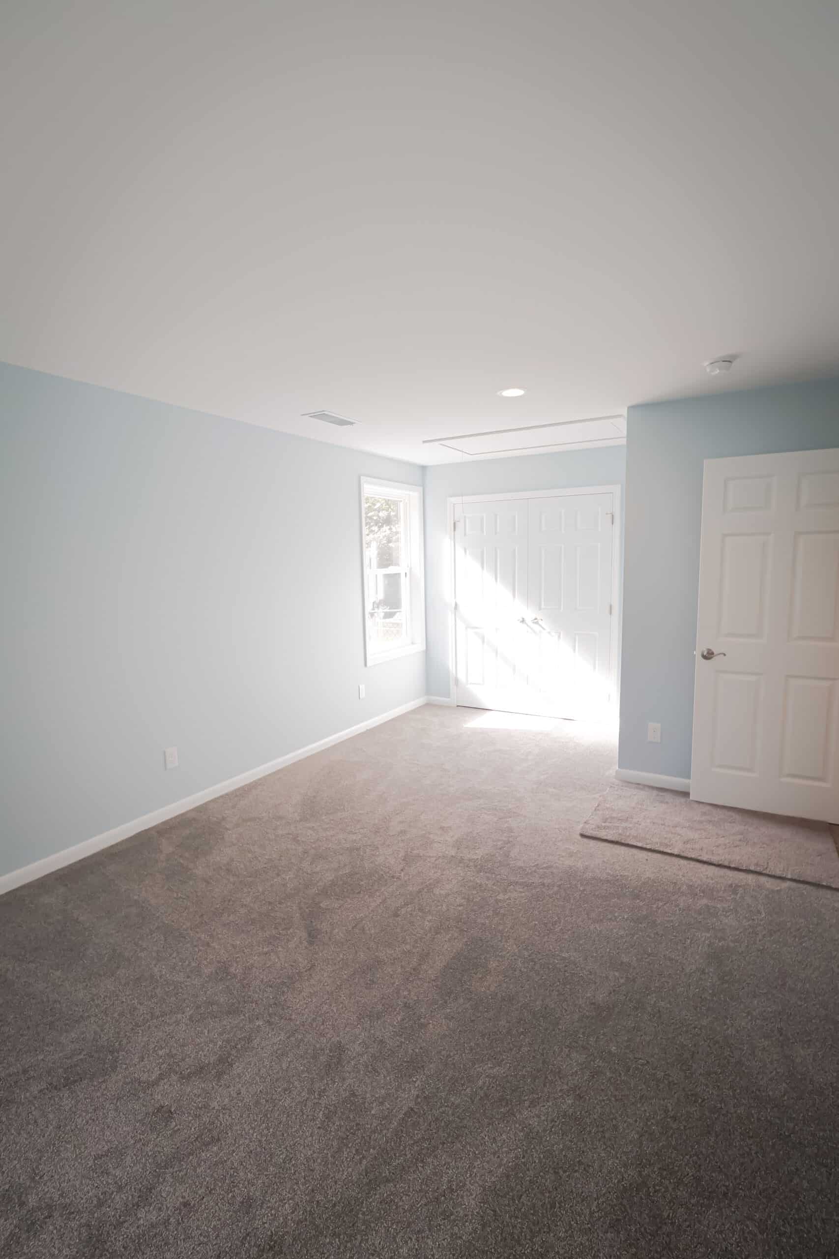 A bright, empty room with light blue walls, gray carpet, a window, and two white doors, one letting in natural sunlight. The space is clean and uncluttered.