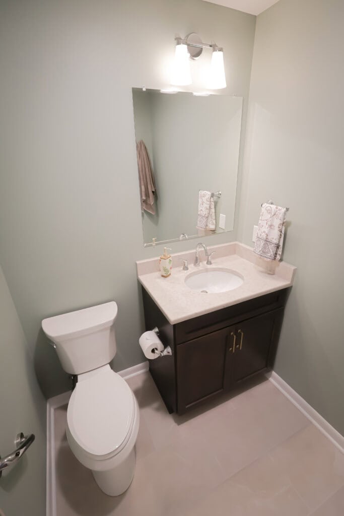 A small, modern bathroom with a white toilet, a dark wood vanity with a white sink, a large mirror, and a light fixture above. Towels and soap are neatly arranged on the countertop. Walls are painted a light gray-green.