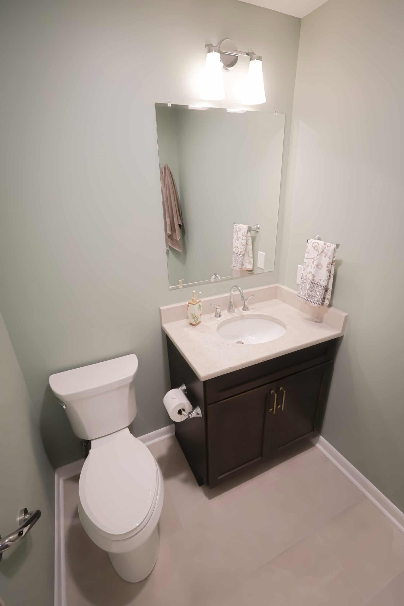 A small, modern bathroom with a white toilet, a dark wood vanity with a white sink, a large mirror, and a light fixture above. Towels and soap are neatly arranged on the countertop. Walls are painted a light gray-green.