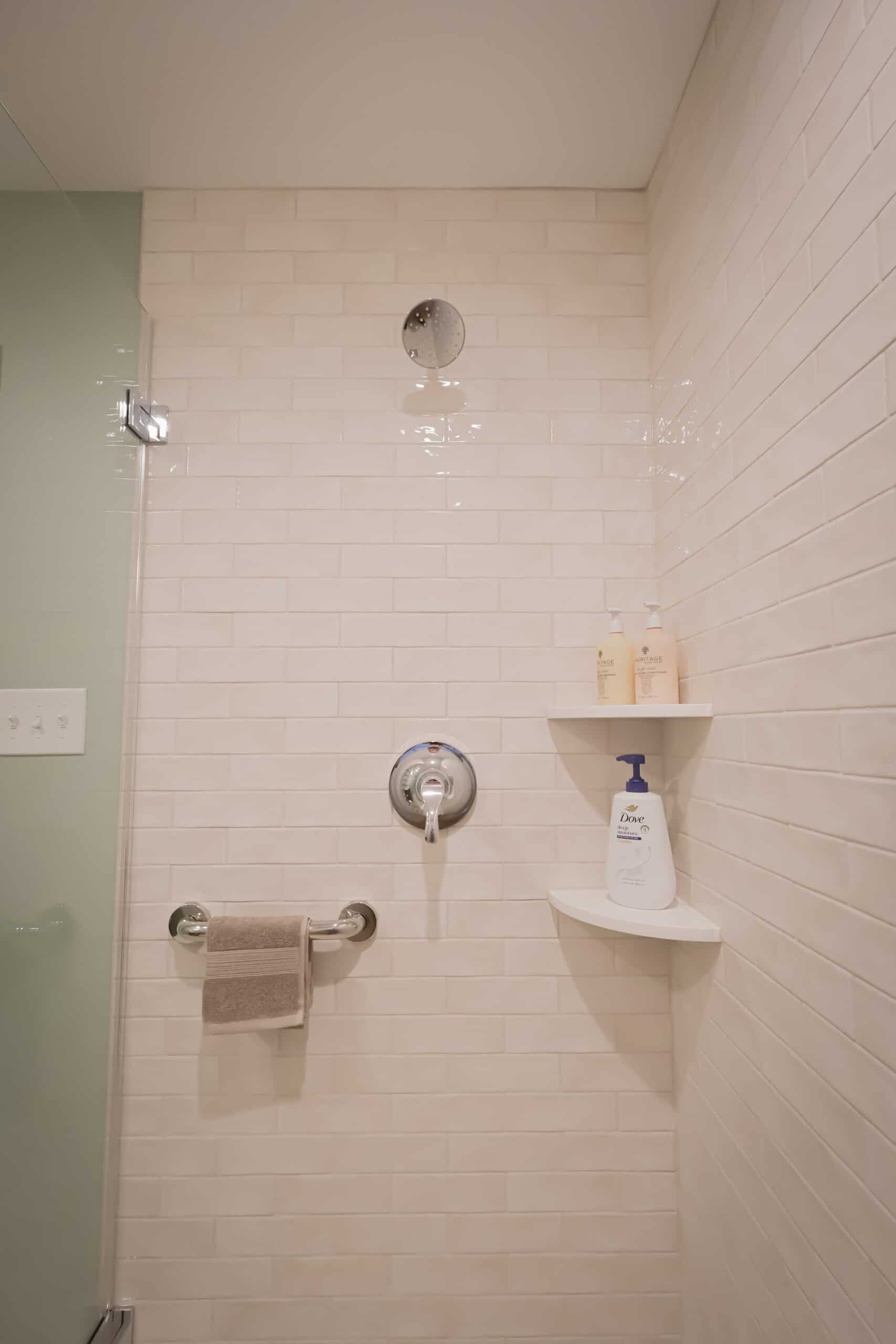 A clean shower with white subway tile walls, a silver shower control, a glass door, a towel on a grab bar, and two corner shelves holding soap bottles and a pump bottle.